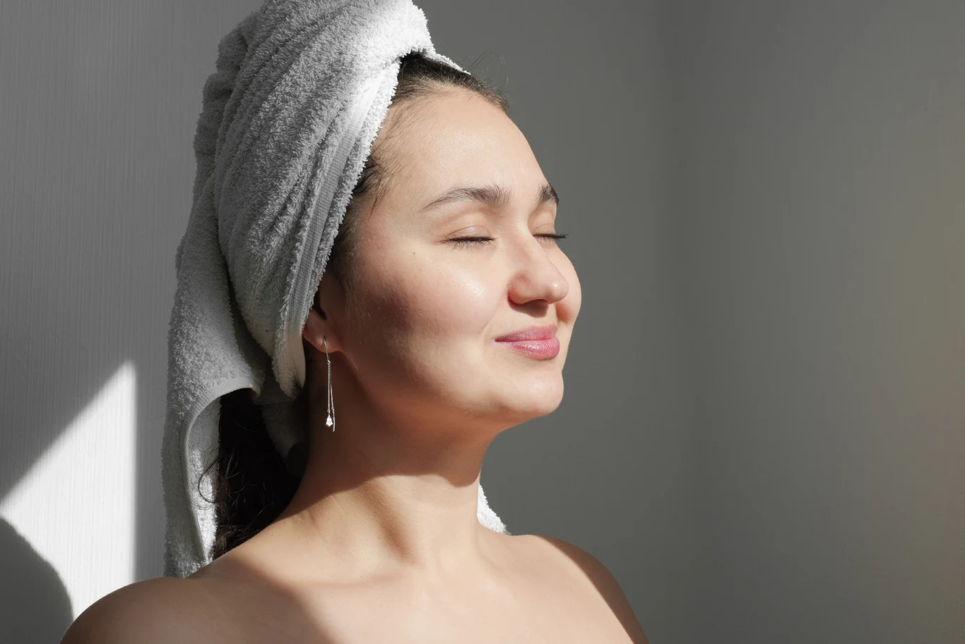 Kobieta z ręcznikiem owiniętym wokół głowy, stojąca w naturalnym świetle z zamkniętymi oczami i delikatnym uśmiechem na twarzy, sprawiająca wrażenie relaksu i odprężenia.