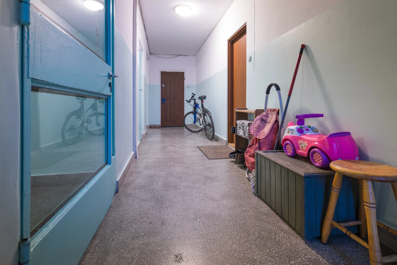 Meble na korytarzach w polskich blokach to częsty widok. Korytarz w budynku mieszkalnym z zaparkowanym rowerem, dziecięcym jeździkiem, różowym wózkiem na zabawki, małym stołkiem oraz szafką na buty. Po lewej stronie przeszklone drzwi, podłoga z szarego materiału.
