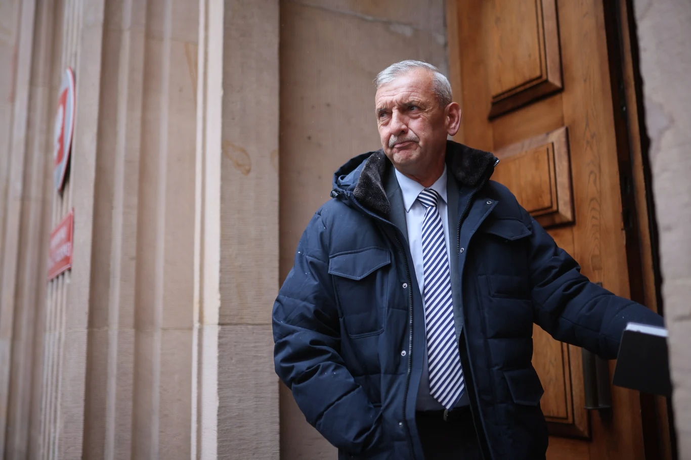 W MEN spotkanie ws. wzrostu wynagrodzeń nauczycieli. Według ZNP propozycja resortu nieakceptowana Starszy mężczyzna w ciemnej kurtce i krawacie stoi przy otwartych drzwiach budynku z kamiennymi ścianami, patrząc w bok z poważnym wyrazem twarzy.