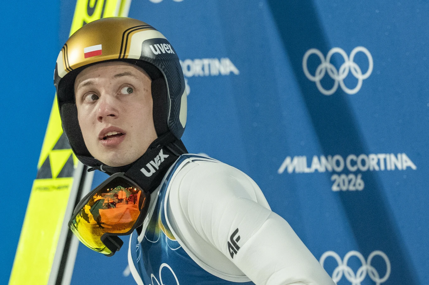 Kacper Tomasiak przeszedł do historii polskiego sportu, jako jeden z czwórki zdobywców trzech medali na jednych igrzyskach Sportowiec w kombinezonie narciarskim i kasku z widoczną polską flagą na tle niebieskiej ścianki z logo igrzysk olimpijskich Milano Cortina 2026.
