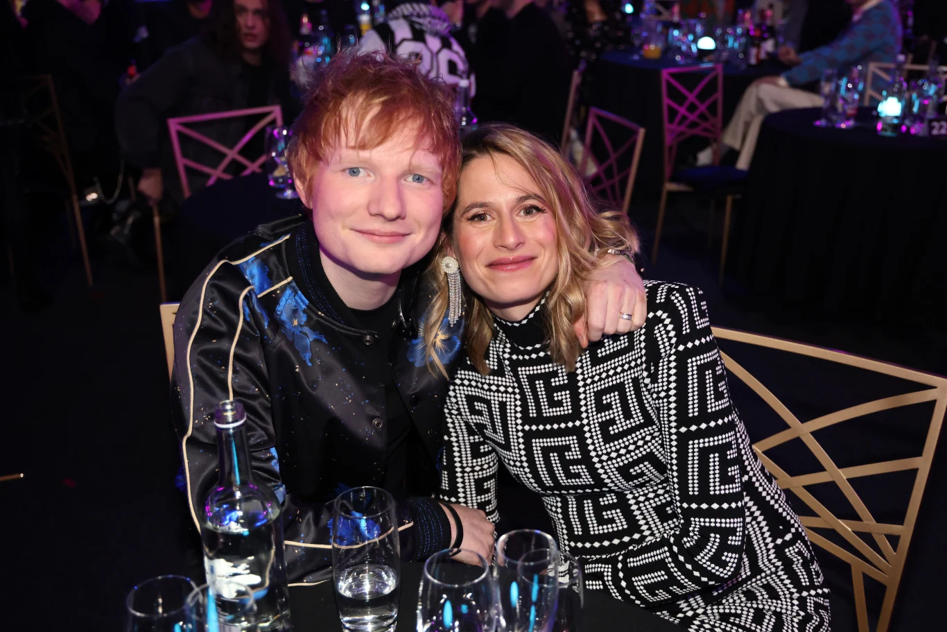Ed Sheeran i jego żona Cherry Seaborn Para siedzi razem przy stoliku na eleganckiej gali, trzymają się blisko siebie i uśmiechają do kamery, na stole butelka wody, kieliszki oraz szklanki, w tle inni uczestnicy wydarzenia.