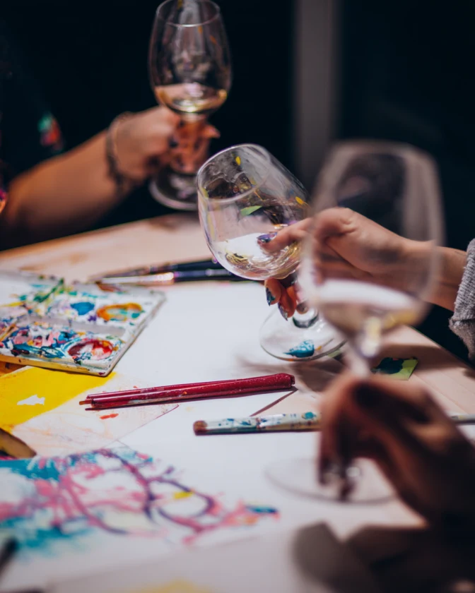 Stół z przyborami do malowania, kieliszki z białym winem w dłoniach, kolorowe farby i kartki papieru, tworząc atmosferę wspólnej kreatywności i relaksu.