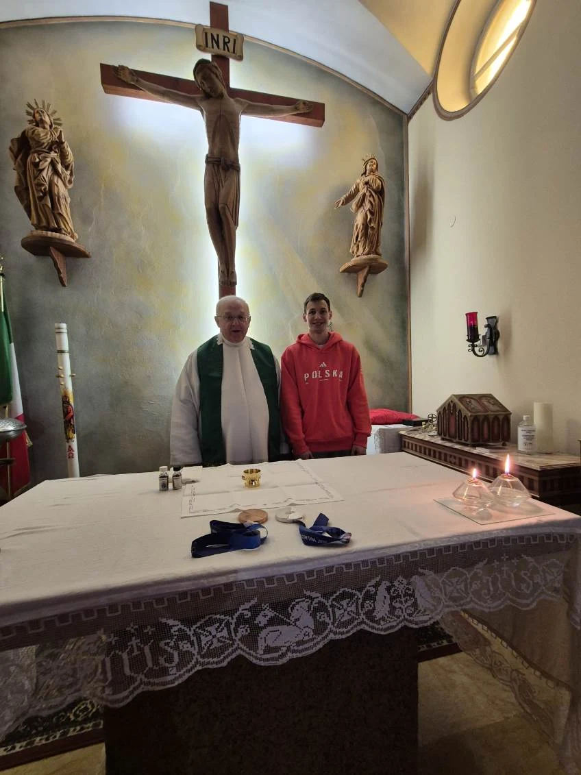 Ksiądz Edward Pleń i Kacper Tomasiak Duchowny w szatach liturgicznych i młody mężczyzna stoją razem przed ołtarzem w kościele, nad nimi duży krucyfiks oraz dwie figury świętych, na ołtarzu ustawione elementy liturgiczne i zapalona świeca