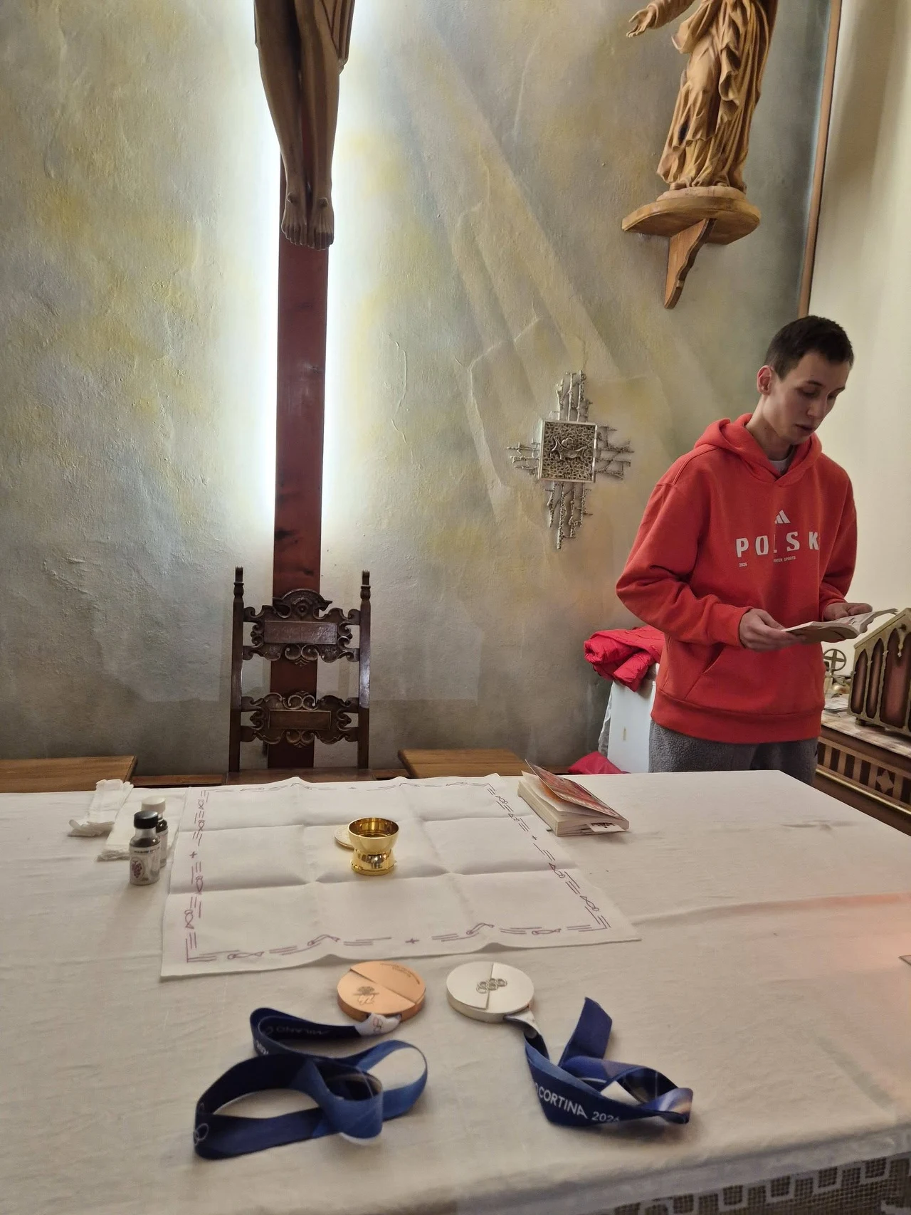 Kacper Tomasiak w roli lektora na mszy świętej w wiosce olimpijskiej Młody mężczyzna w czerwonej bluzie z napisem Polska stoi przy ołtarzu w kościele, na ołtarzu leżą medale sportowe, kielich, hostie oraz inne przedmioty liturgiczne.