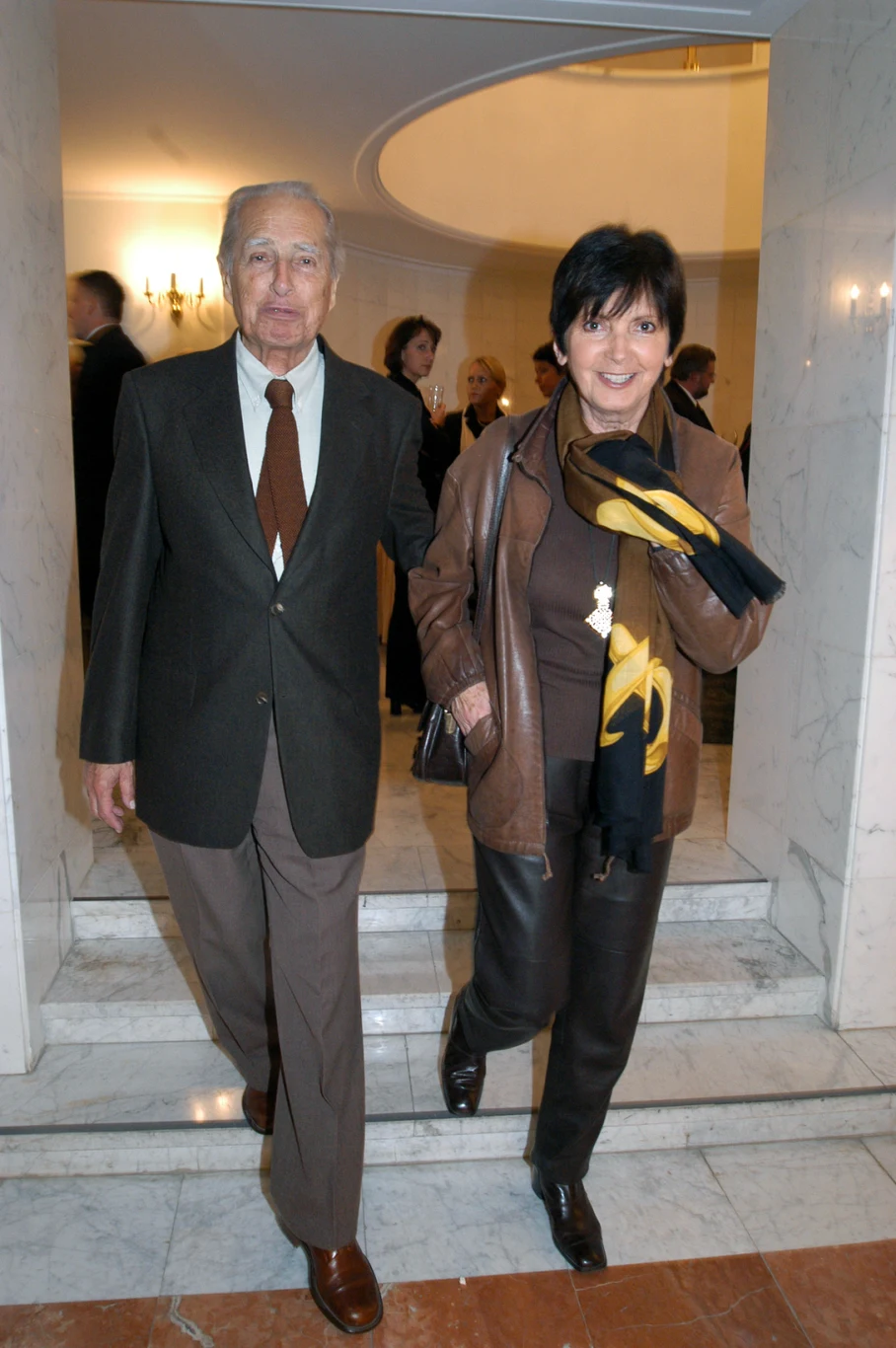 Elegancka para w średnim wieku wchodząca po marmurowych schodach w jasnym, reprezentacyjnym wnętrzu. Kobieta ubrana w ciemnobrązową kurtkę i szalik w żółtym kolorze, mężczyzna w garniturze z brązowym krawatem. Za nimi grupa osób w tle.