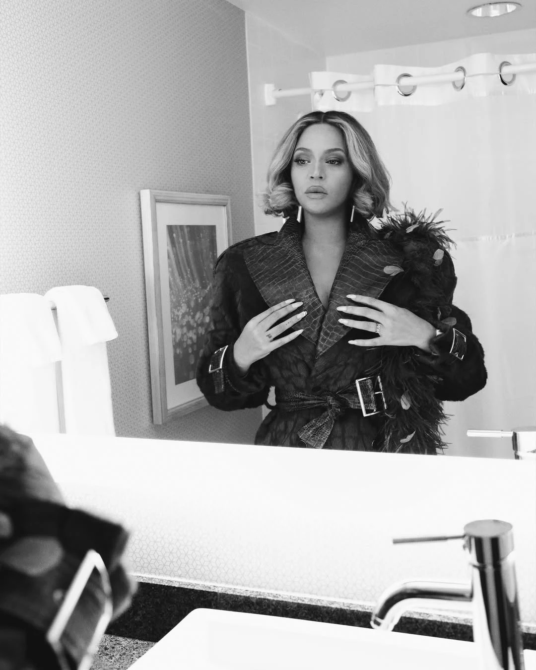 Kobieta ubrana w elegancki płaszcz z futrzanymi akcentami stoi przed lustrem w łazience, patrząc na swoje odbicie z powagą. W tle widoczne są elementy wyposażenia łazienki, takie jak ręczniki i zasłona prysznicowa.