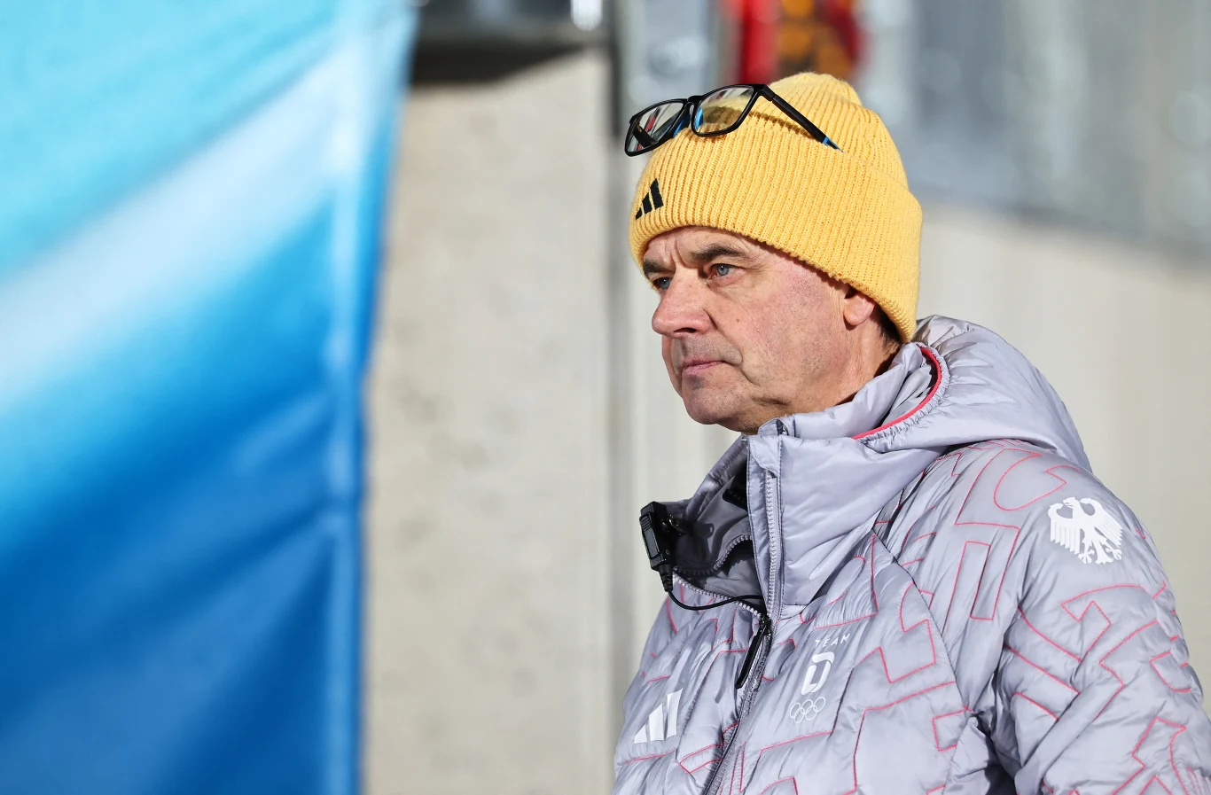 Mężczyzna w żółtej czapce i okularach na czubku głowy, ubrany w zimową kurtkę, stoi na zewnątrz i patrzy w jednym kierunku, na tle rozmytej niebieskiej i szarej scenerii.