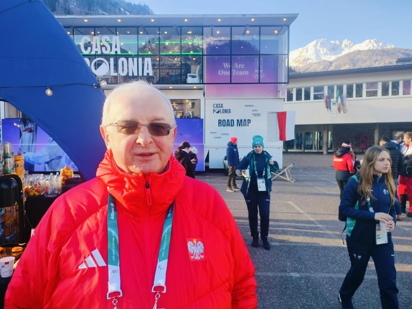 Edward Pleń Starszy mężczyzna w czerwonej kurtce z polskim godłem, z zawieszoną akredytacją, stoi na tle nowoczesnego budynku Casa Polonia, wokół kręci się kilka osób, a w tle widoczne są ośnieżone góry.