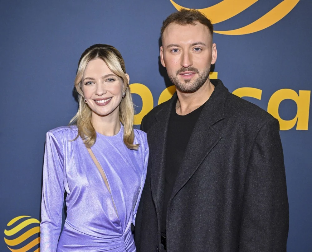 Paulina Gałązka i Michał Bartkiewicz Uśmiechnięta kobieta w jasnofioletowej sukni oraz mężczyzna w ciemnym płaszczu stoją razem obok siebie na tle reklamowym z żółtym napisem i logotypem.