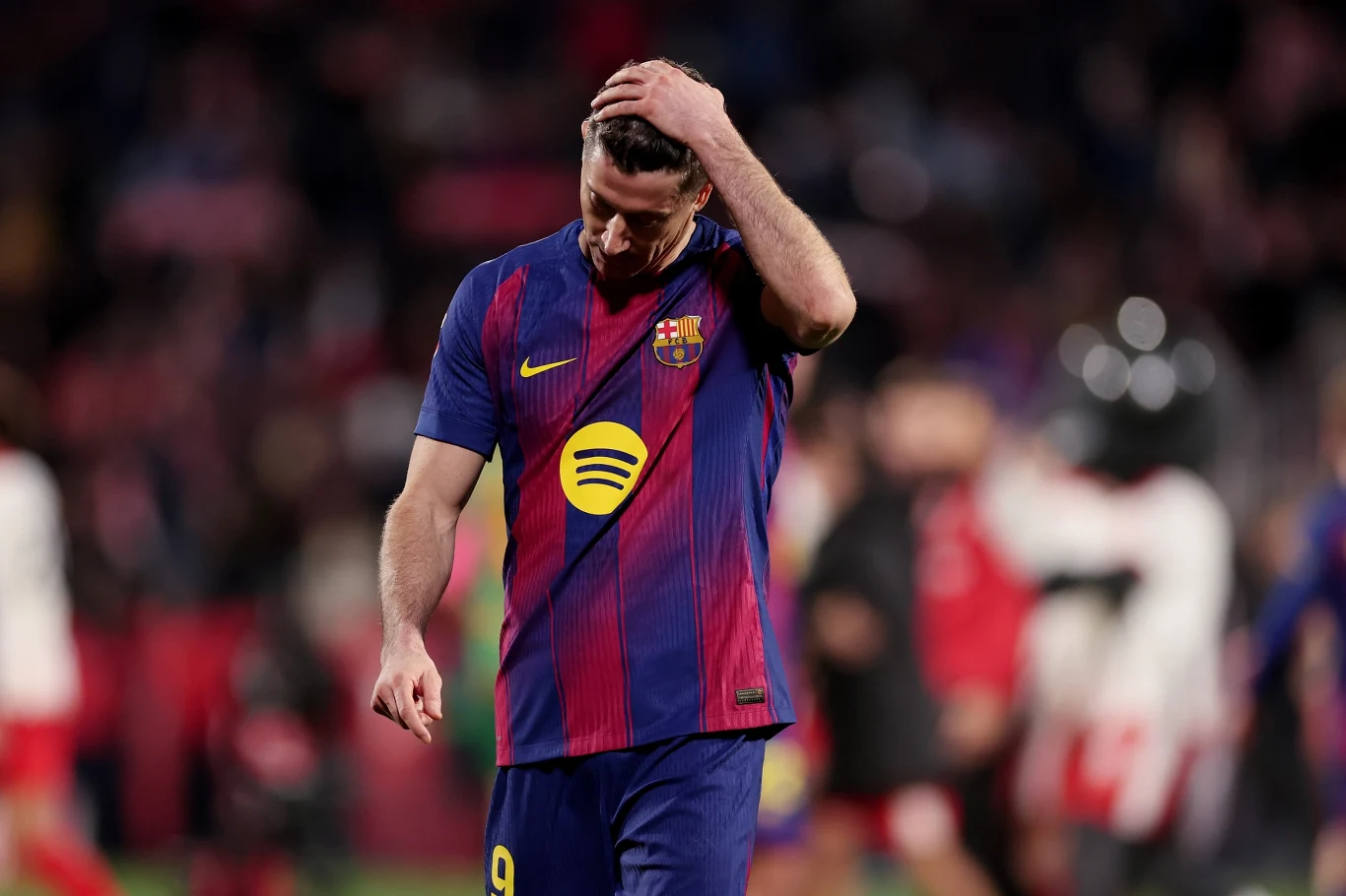 Robert Lewandowski Piłkarz w barwach FC Barcelony ze spuszczoną głową i ręką na głowie sugeruje rozczarowanie po meczu, w tle rozmyta publiczność oraz inni zawodnicy.