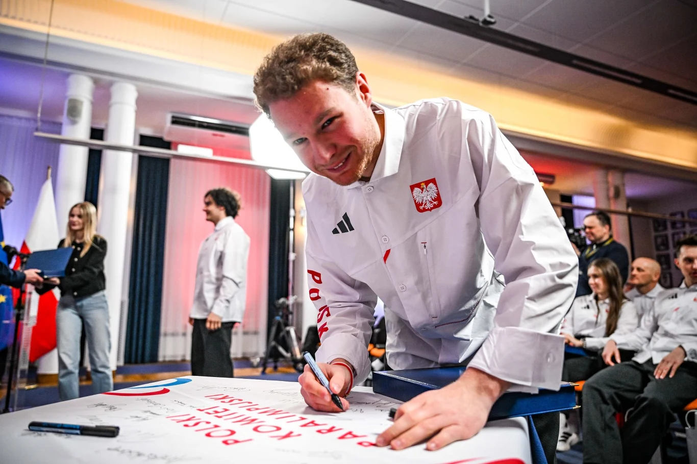 Mężczyzna w białej sportowej kurtce z godłem Polski podpisuje duży plakat przy stole, w tle kilku innych uczestników wydarzenia w podobnych strojach, a wokół nich panuje przyjazna atmosfera spotkania.