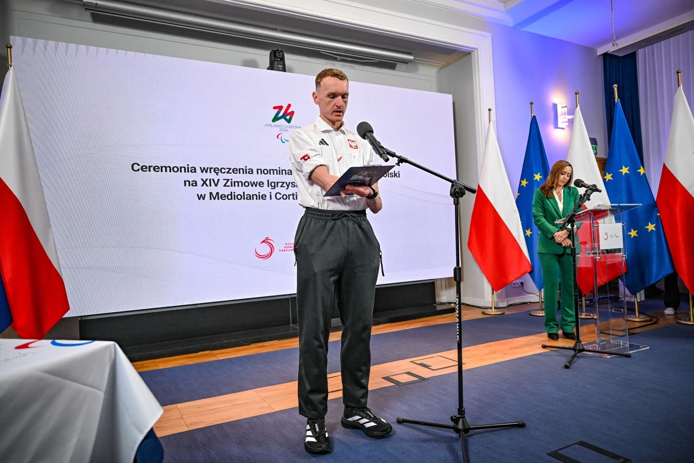 Mężczyzna w sportowym stroju reprezentacyjnym przemawia przy mikrofonie na tle ekranu z napisem dotyczącym ceremonii wręczenia nominacji olimpijskich, w tle widoczne są polskie i unijne flagi oraz kobieta przy mównicy.
