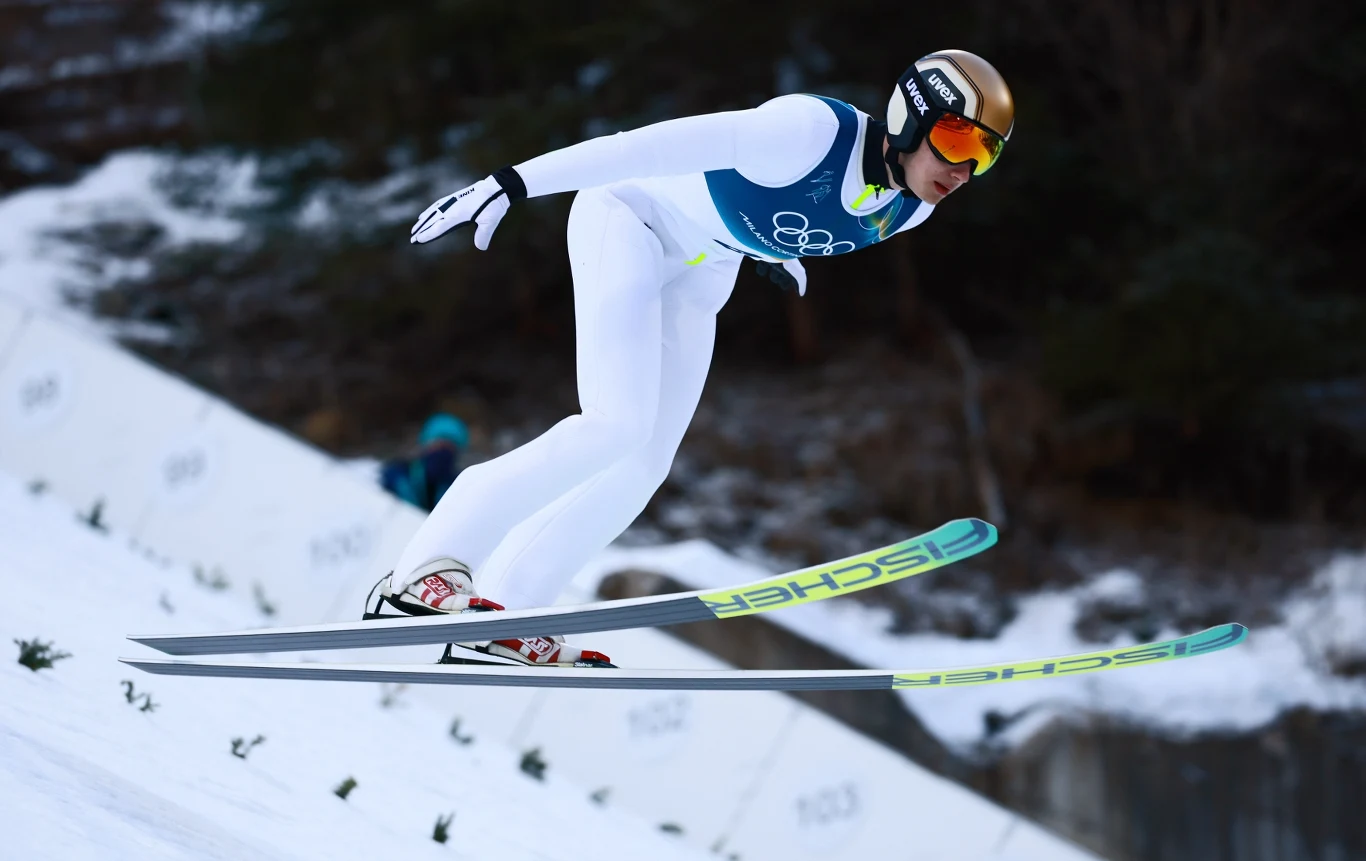 Zawodnik w kombinezonie do skoków narciarskich w trakcie lotu na skoczni, w tle śnieżne zbocze oraz rozmyte drzewa.