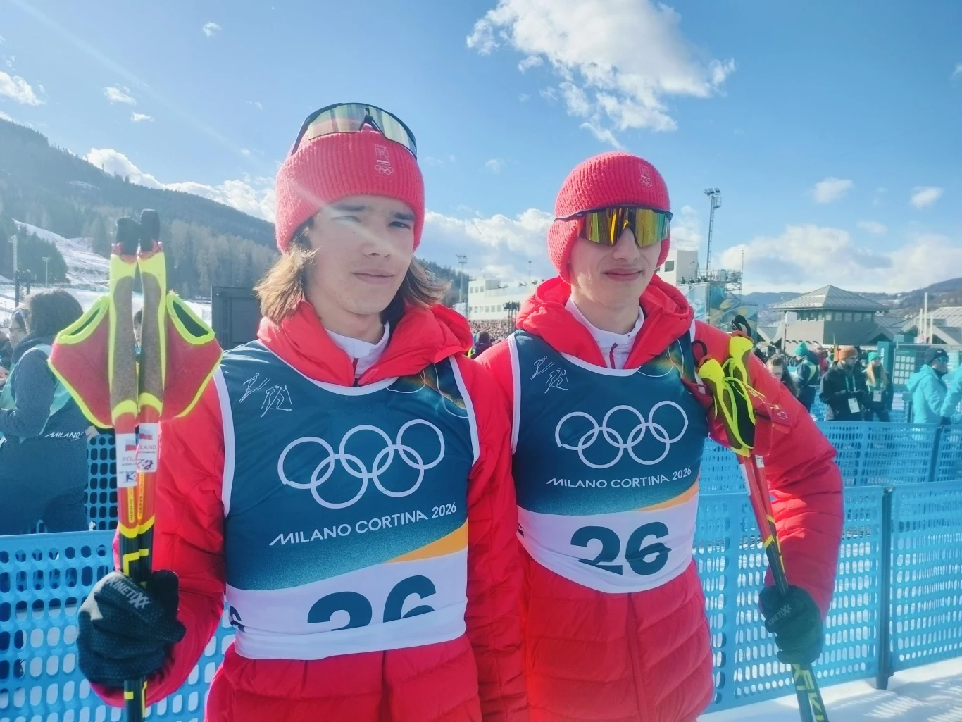 Dwóch młodych sportowców w czerwonych kombinezonach i czapkach, trzymających kijki narciarskie, stoi obok siebie na tle zimowego krajobrazu i infrastruktury zawodów olimpijskich. Na ich strojach widoczne są numery startowe z logo igrzysk olimpijskich M...