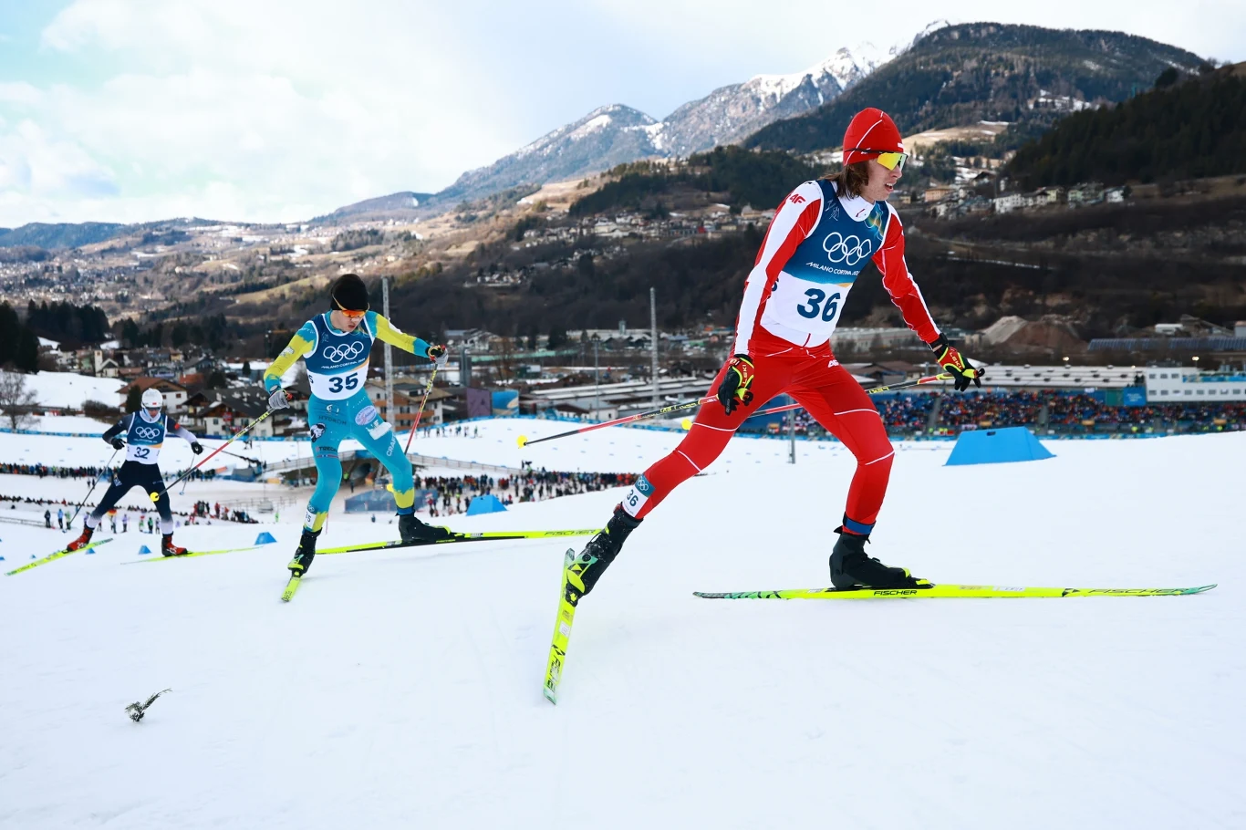 Trzech biegaczy narciarskich rywalizujących na śnieżnej trasie na tle górskich szczytów i zimowego krajobrazu, wszyscy ubrani w sportowe stroje z numerami startowymi oraz charakterystycznymi nartami biegowymi.