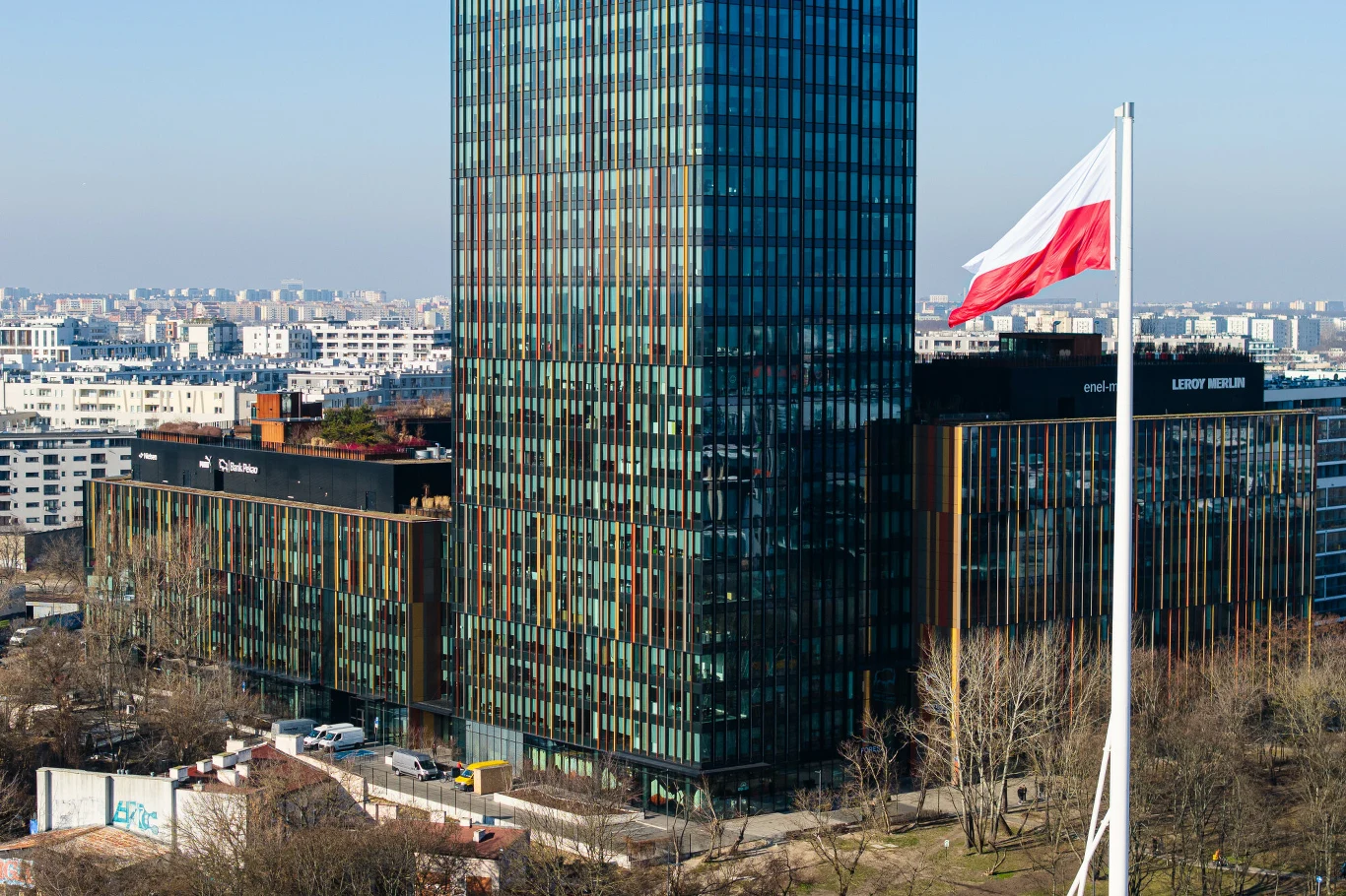 Brytyjski ekonomista chwali polską gospodarkę. Mówi, że polska bardzo się zmieniła Wysoki, nowoczesny biurowiec ze szklaną fasadą, na pierwszym planie biało-czerwona flaga Polski na masztcie. W tle rozległa panorama miasta z wieloma białymi budynkami mieszkalnymi.