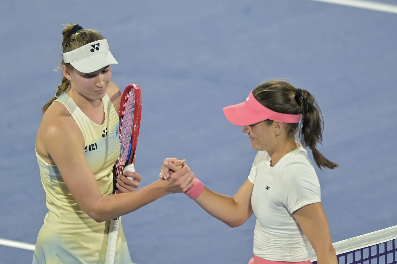 Jelena Rybakina i Kimberly Birrell Dwie tenisistki stoją przy siatce na korcie, podają sobie dłonie w geście sportowego szacunku po zakończonym meczu. Jedna z zawodniczek trzyma rakietę, obie ubrane w sportowe stroje oraz daszki, w tle widoczny niebieski kort.