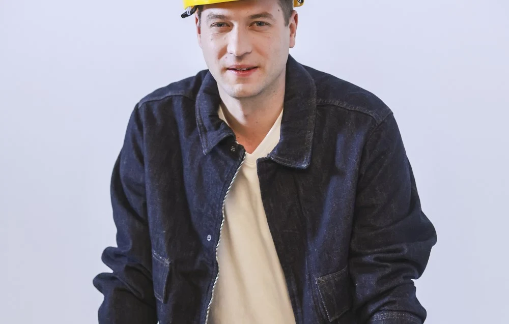 Mężczyzna w żółtym kasku ochronnym i ciemnej kurtce roboczej stoi na tle jasnego tła, patrząc w stronę aparatu