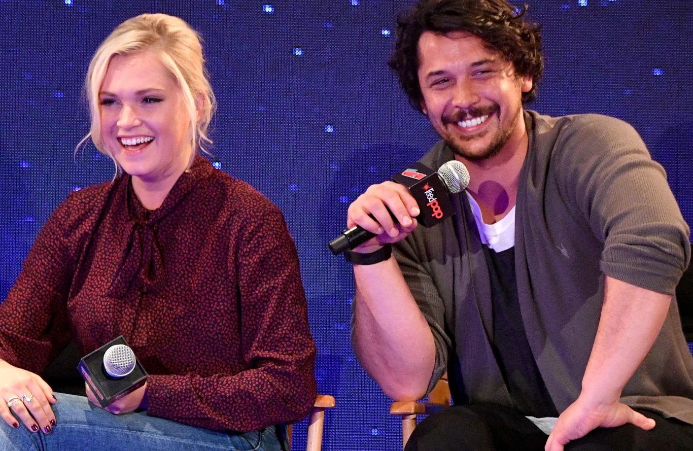 Eliza Taylor i Bob Morley podczas New York Comic Con Dwie uśmiechnięte osoby siedzą na scenie, trzymając mikrofony, w tle granatowa powierzchnia z jasnymi punktami przypominającymi gwiazdy.
