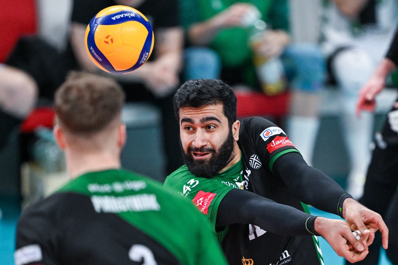 Zawodnik siatkarski skupiony na odbiorze piłki podczas meczu, w tle widoczny inny gracz tej samej drużyny, obaj ubrani w zielono-czarne stroje sportowe.