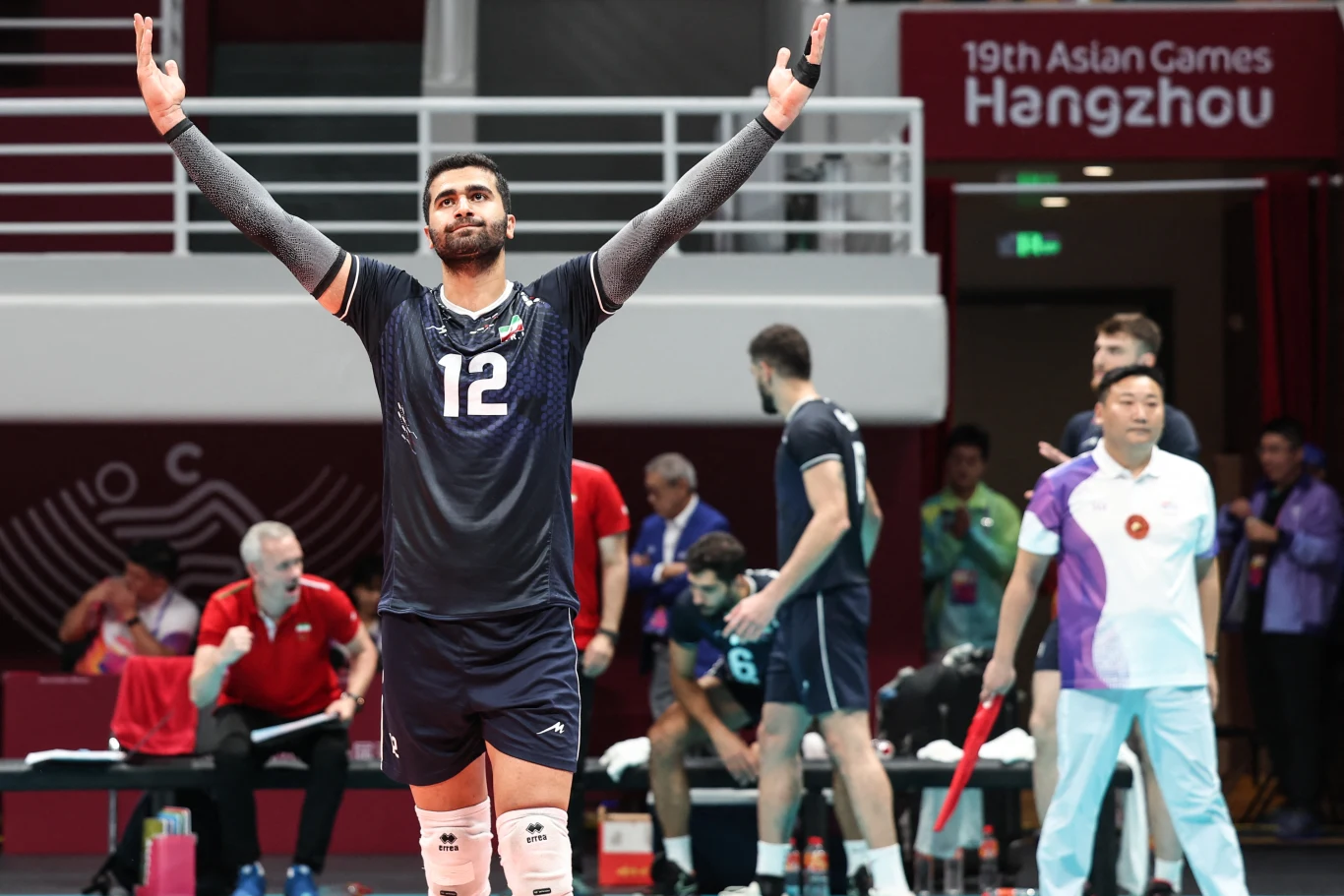 Zawodnik w ciemnym stroju z numerem 12 unosi ręce w górę podczas meczu siatkówki, w tle widoczni są inni gracze, sędziowie oraz baner 19. Asian Games Hangzhou.