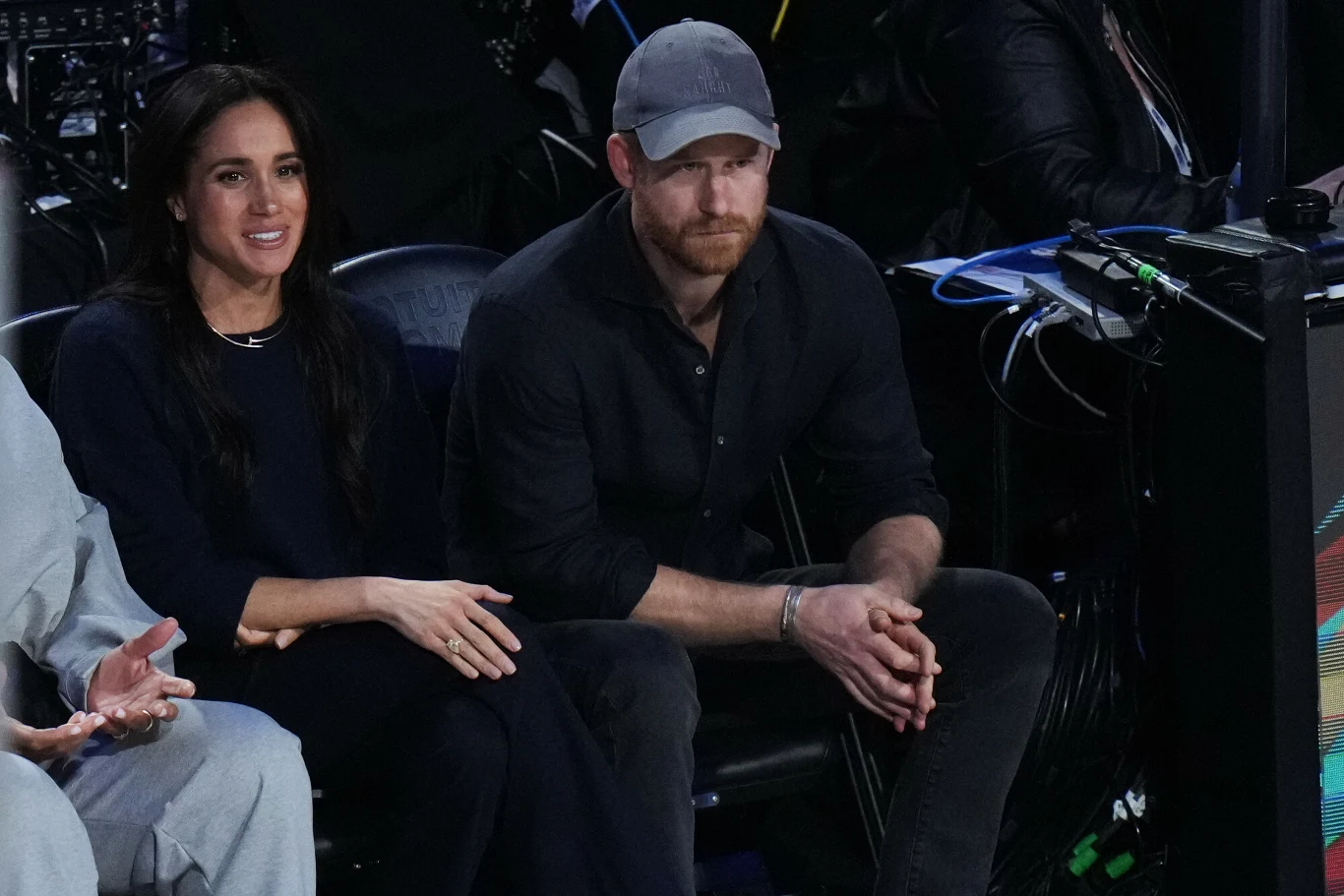 Para siedząca razem podczas wydarzenia sportowego, mężczyzna w szarej czapce i ciemnej koszuli, kobieta w ciemnym stroju z długimi włosami, oboje skoncentrowani na tym, co dzieje się przed nimi, na pierwszym planie widoczne dłonie innej osoby.