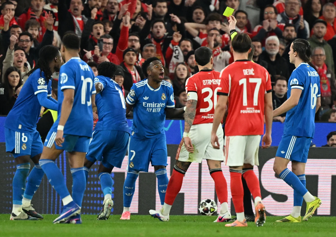 Sędzia pokazuje żółtą kartkę zawodnikowi drużyny w czerwonych koszulkach otoczonemu przez piłkarzy drużyny w niebieskich strojach, na tle pełnych emocji kibiców na trybunach.