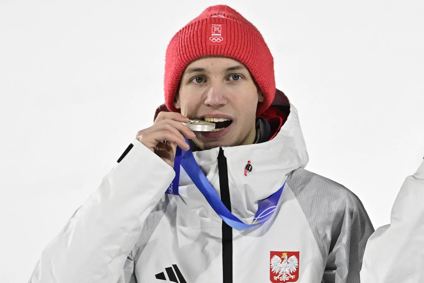 Sportowiec w białej kurtce z biało-czerwonym godłem Polski oraz czerwonej czapce gryzie srebrny medal zawieszony na niebieskiej wstążce, wyrażając radość z osiągnięcia.
