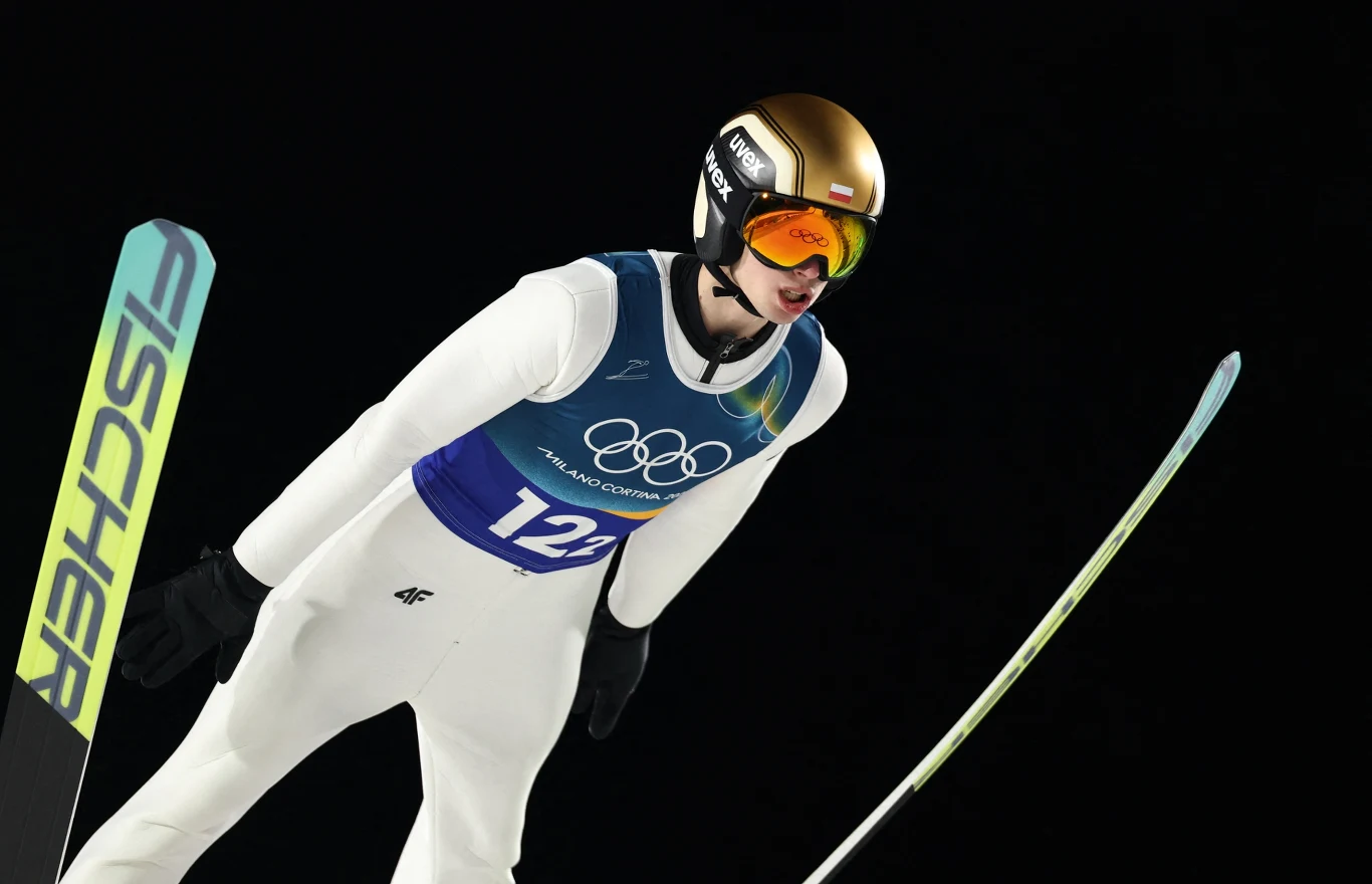 Skoczek narciarski w kombinezonie sportowym i kasku w trakcie lotu podczas zawodów, na piersi numer startowy 12, w tle ciemne niebo, widoczne narty i sportowa determinacja zawodnika.