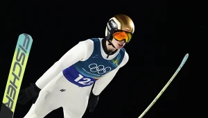 Skoczek narciarski w kombinezonie sportowym i kasku w trakcie lotu podczas zawodów, na piersi numer startowy 12, w tle ciemne niebo, widoczne narty i sportowa determinacja zawodnika.