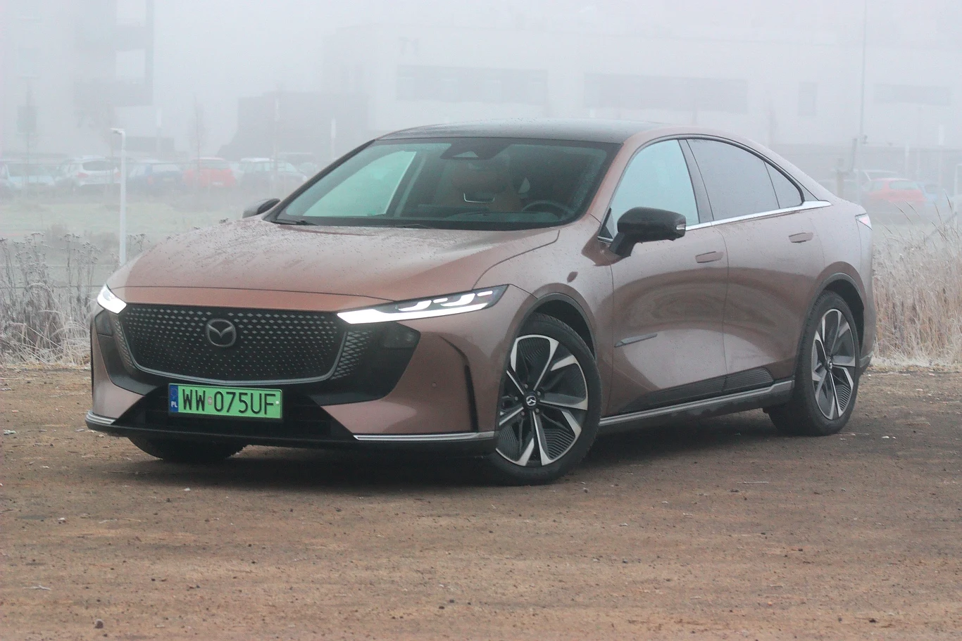 Mazda 6e to samochód oparty na modelu chińskiej firmy Changan Brązowy samochód Mazda o nowoczesnym, aerodynamicznym kształcie stoi na nieutwardzonym podłożu, otoczony lekką mgłą ze słabo widocznymi budynkami w tle, auto posiada zielone tablice rejestracyjne.