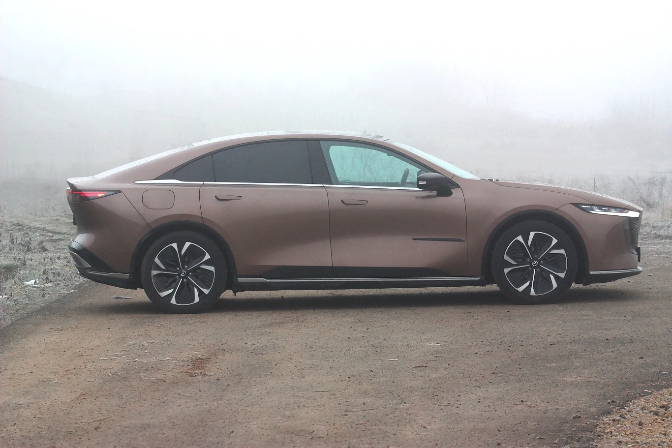 Mazda 6e naprawdę wygląda jak model 6 nowej generacji Brązowy luksusowy sedan elektryczny zaparkowany na szutrowej drodze, otoczony mglistym, pustynnym krajobrazem, nowoczesny design samochodu wyróżniają aerodynamiczne linie i charakterystyczne felgi.