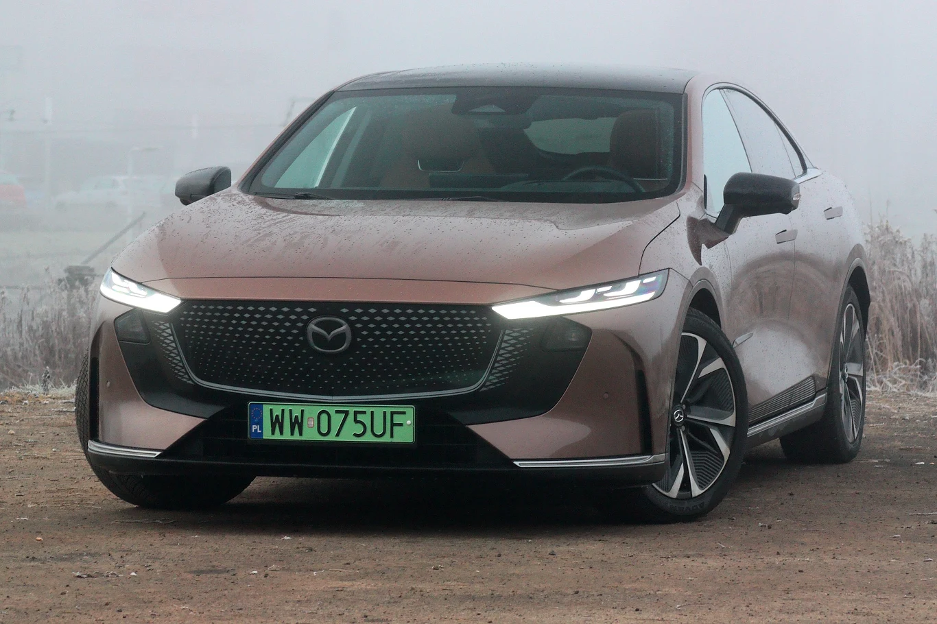 Mazda 6e waży niespełna dwie tony Brązowy samochód osobowy Mazda stoi na nieutwardzonej drodze w otoczeniu mgły, z charakterystycznym przednim grillem oraz włączonymi światłami dziennymi.
