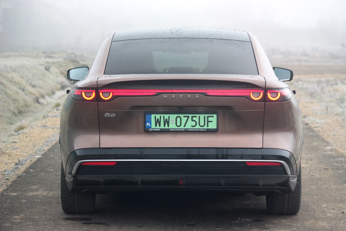 Mazda 6e ma dwie wersje napędowe, a wybór wcale nie jest oczywisty Nowoczesny samochód osobowy w kolorze brązowym stoi na drodze, widok od tyłu, z widoczną zieloną tablicą rejestracyjną, światłami i logo marki; otoczenie jest zamglone, z lekkim szronem pobocza.