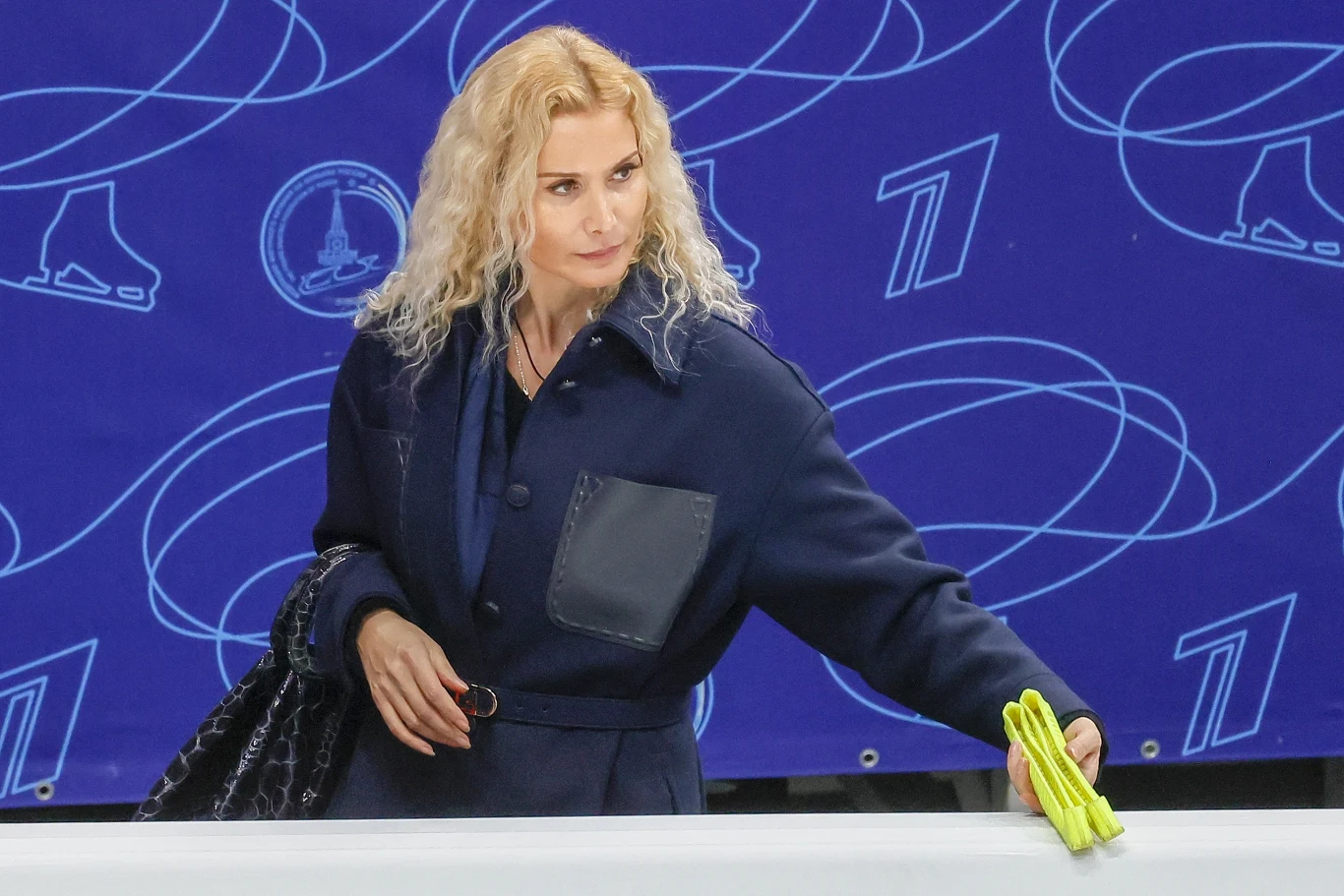 Kobieta o blond, kręconych włosach w granatowym płaszczu trzymająca żółtą szmatkę, stoi przy barierce na tle niebieskiej ściany z grafiką zawierającą łyżwy oraz numer 1.