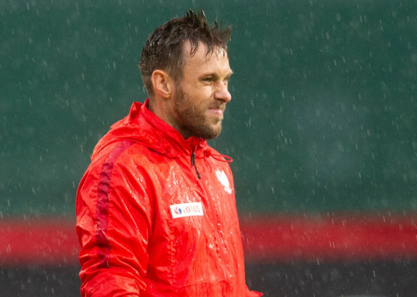 Mężczyzna w czerwonej kurtce sportowej stoi na zewnątrz podczas deszczu, z widocznymi kroplami wody i zielonym rozmytym tłem.