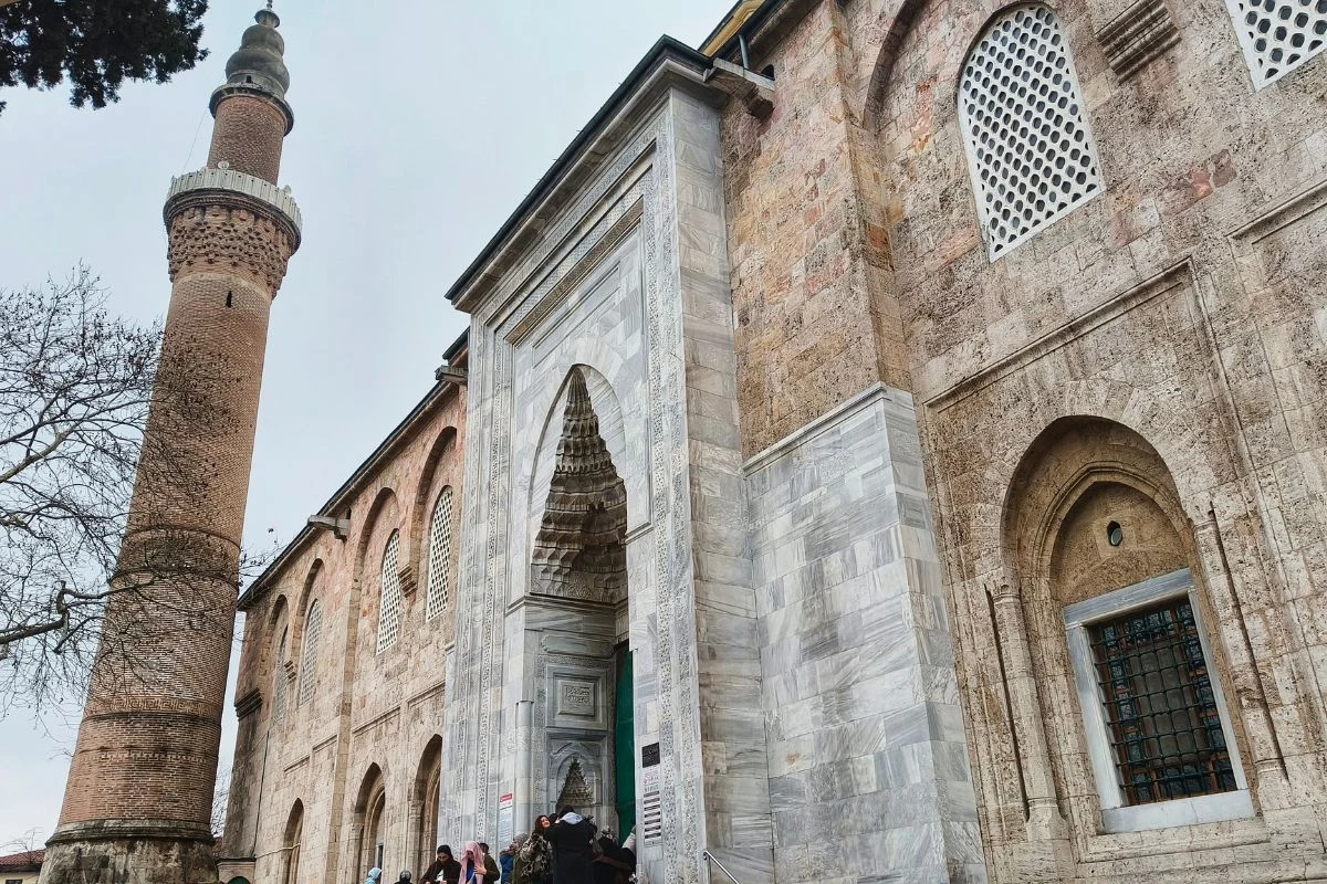 Wielki Meczet to jeden z najważniejszych zabytków Bursy Kamienna fasada zabytkowego meczetu z wysokim minaretem, duży ozdobny portal wejściowy, liczne łuki oraz detale architektoniczne typowe dla stylu islamskiego. Przed wejściem gromadzi się grupa ludzi, obok rośnie drzewo.