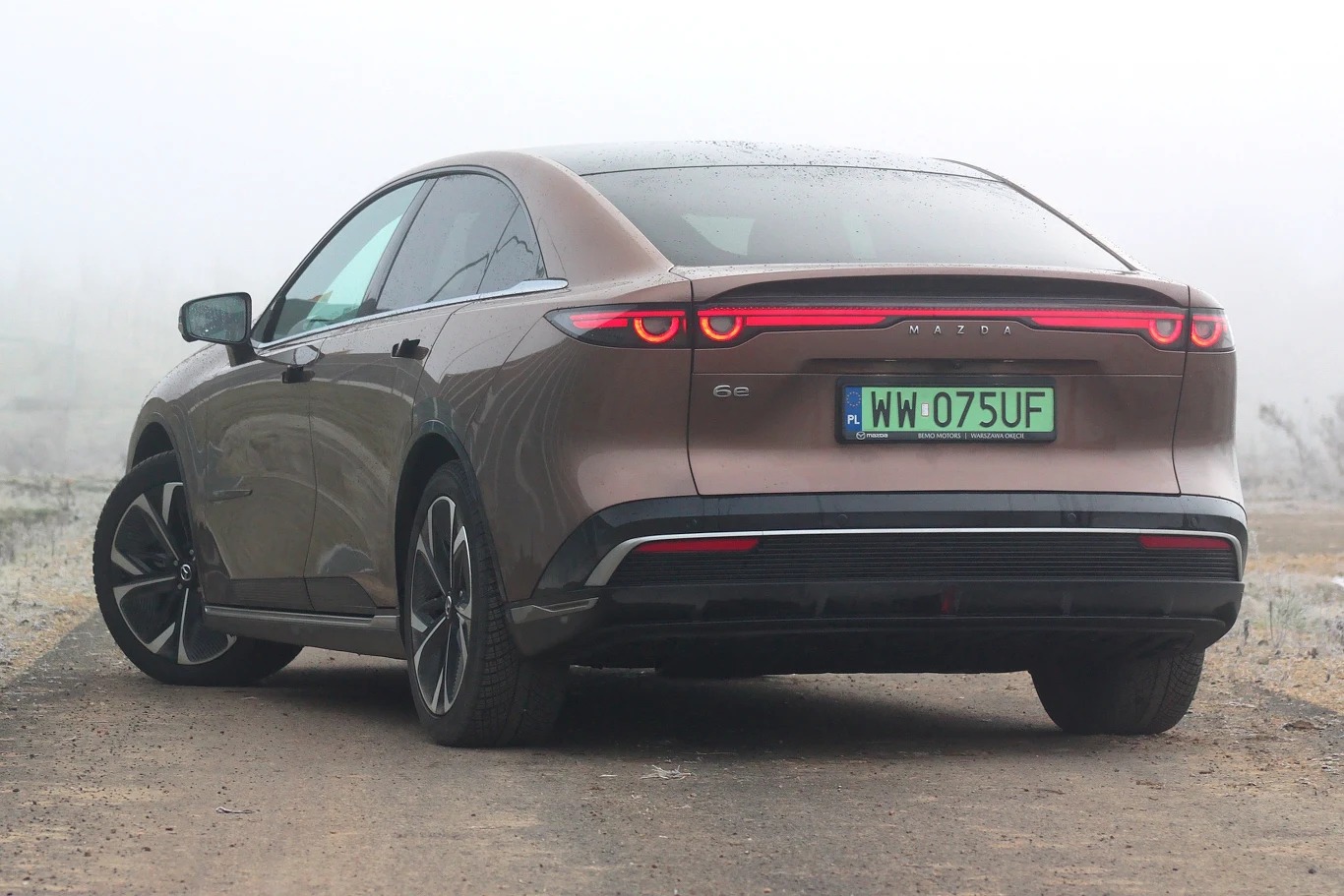 Mazda 6e jest oferowana w dwóch wersjach wyposażeniowych. Już bazowa ma niemal wszystko Brązowy sedan elektryczny marki Nio ET7 sfotografowany z tyłu na drodze w pochmurny dzień, zielona tablica rejestracyjna, nowoczesny design świateł tylnych oraz aerodynamiczna sylwetka nadwozia.