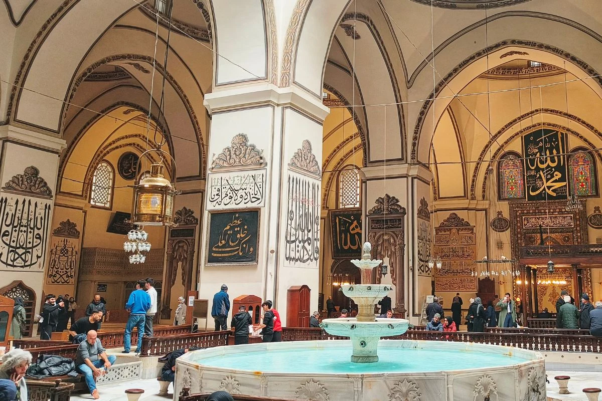 Bursa to dobre miejsce, by zobaczyć codzienne życie mieszkańców Wnętrze dużego meczetu z centralnie umieszczoną fontanną, otoczoną ludźmi; na ścianach liczne arabskie napisy kaligraficzne i dekoracje, wysokie łuki oraz ozdobne lampy podkreślają sakralny charakter miejsca.