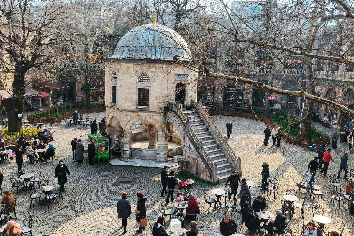 Krótka przerwa podczas zakupów? Koniecznie nad filiżanką tureckiej kawy Dziedziniec z kamiennym pawilonem z kopułą pośrodku, otoczony przez ludzi siedzących przy kawiarnianych stolikach, drzewa bez liści i brukowaną nawierzchnię, luźna atmosfera spotkań i relaksu.