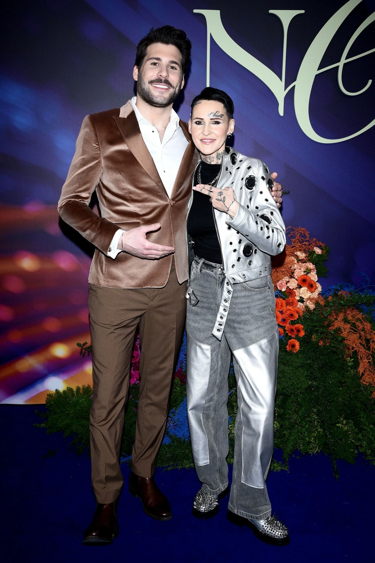 Janek Pirowski i Agnieszka Chylińska chętnie pozowali razem na ściance podczas ramówki TVN. Agnieszka nawet czule objęła kolegę z "Mam talent", a z jej twarzy nie schodził uśmiech. Widać, że bardzo się lubią! Dwoje ludzi pozujących razem na tle ozdobnym, elegancko ubranych, kobieta ma srebrną kurtkę z czarnymi wzorami oraz srebrne buty, mężczyzna ma brązową marynarkę i spodnie, w tle widoczne są kwiaty oraz połyskujący, kolorowy napis.