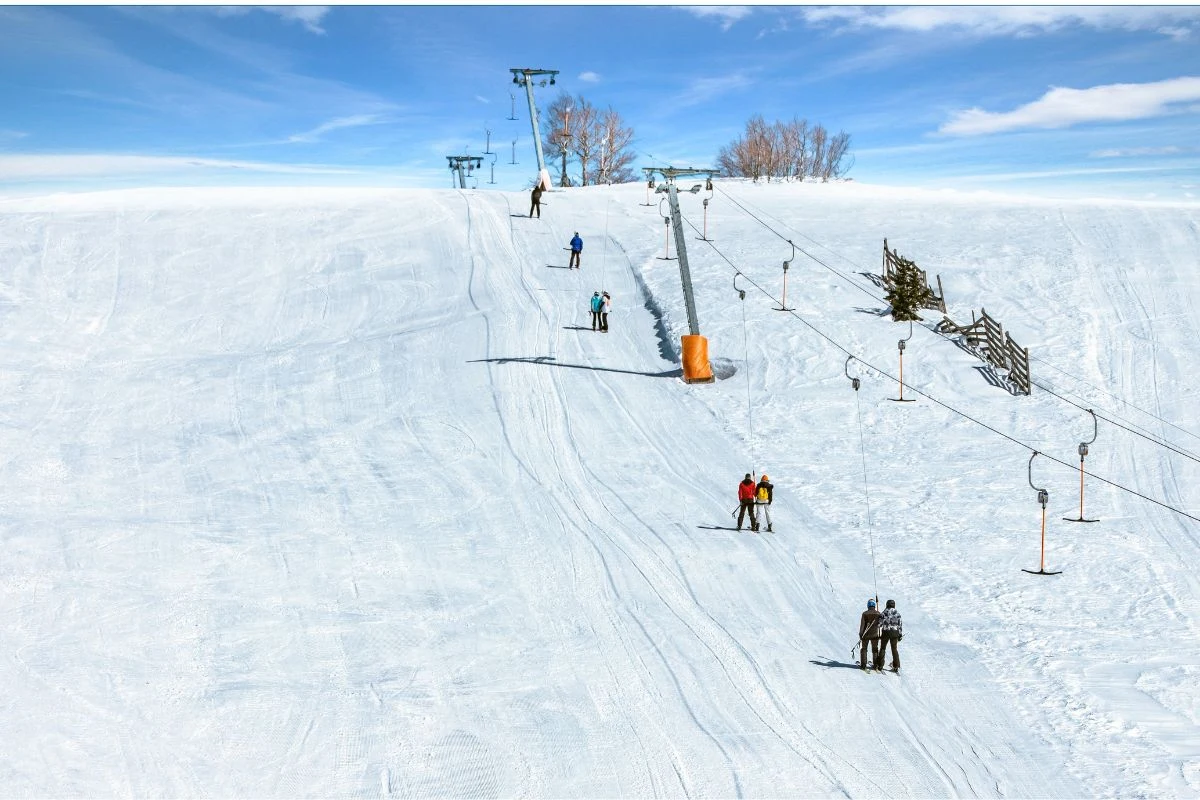 Czy zimowe wakacje w Turcji to dobry pomysł? Jak najbardziej! Grupa narciarzy korzystająca z wyciągu orczykowego na łagodnym stoku pokrytym śniegiem, jasne niebo i niewielkie drzewa na szczycie wzniesienia.