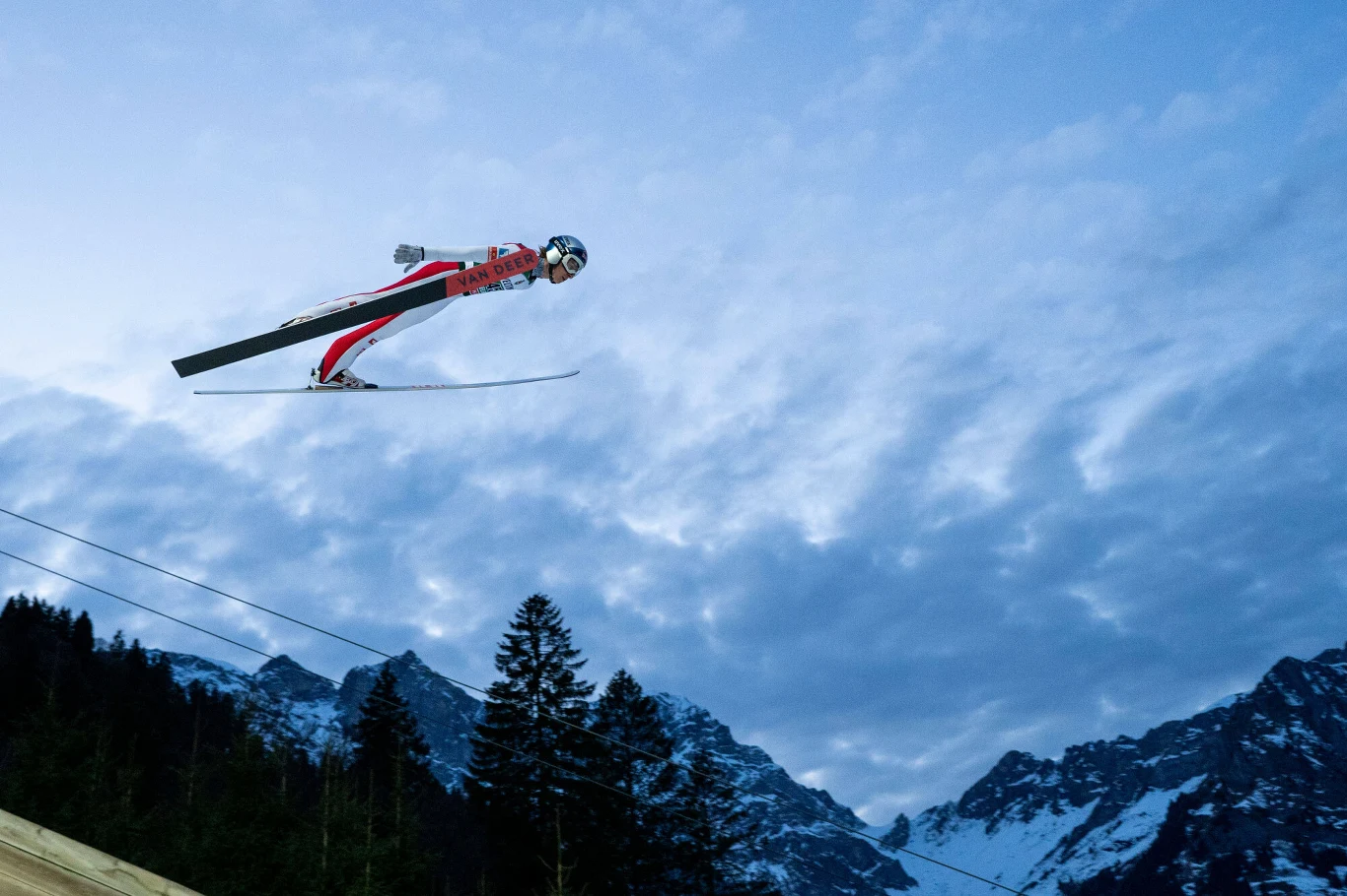 Gross-Titlis-Schanze, Daniel Tschofenig, grudzień 2025 r. Skoczek narciarski unoszący się w powietrzu na tle górskich szczytów pokrytych śniegiem oraz wieczornego nieba, poniżej widoczne są ciemne sylwetki drzew.