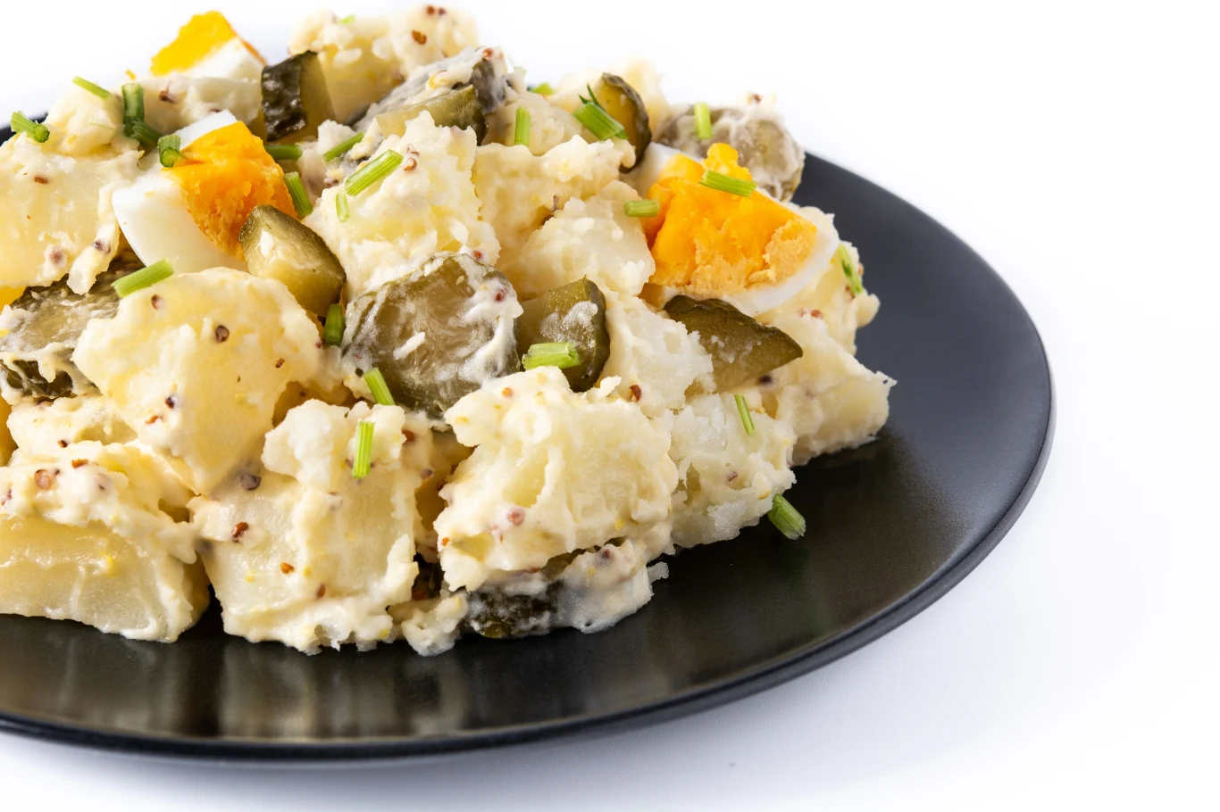 Sałatka z ugotowanych ziemniaków z kawałkami ogórka kiszonego, jajka na twardo i szczypiorku, wszystko wymieszane z musztardą i delikatnym sosem, podane na czarnym talerzu na białym tle.