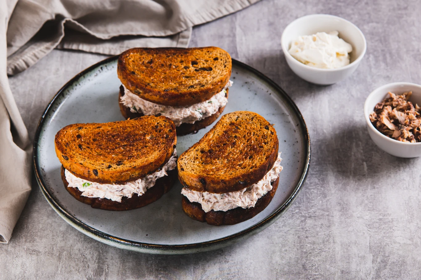 Trzy kanapki z chrupiącego, podsmażanego chleba z kremowym nadzieniem z pasty, podane na talerzu; obok dwie małe miseczki, jedna z kremowym serkiem lub majonezem, druga z pastą rybną lub mięsną.