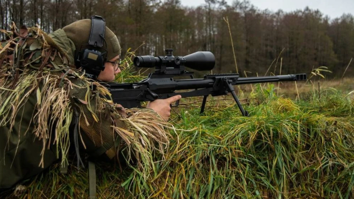 Polska broń ruszyła na podbój rynku. Jest tajemniczy klient Żołnierz w stroju maskującym leży na trawie, celując przez lunetę karabinu snajperskiego, za nim widoczny jest jesienny, zalesiony teren.