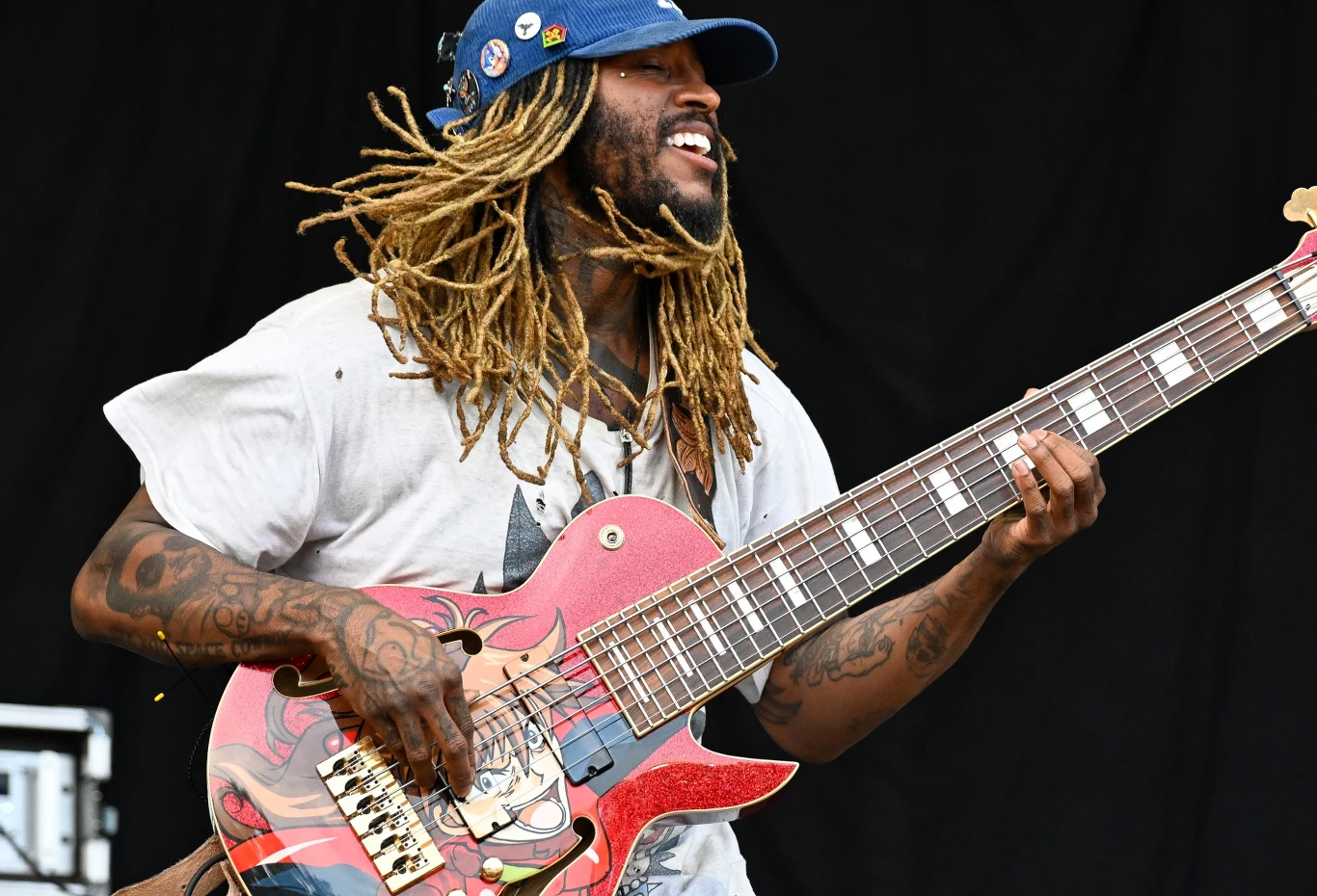 Thundercat zapowiada nowy album Mężczyzna z dredami grający na dużej gitarze basowej ozdobionej kolorową grafiką, uśmiechnięty i ubrany w niebieską czapkę z daszkiem oraz koszulkę, na ciemnym tle.