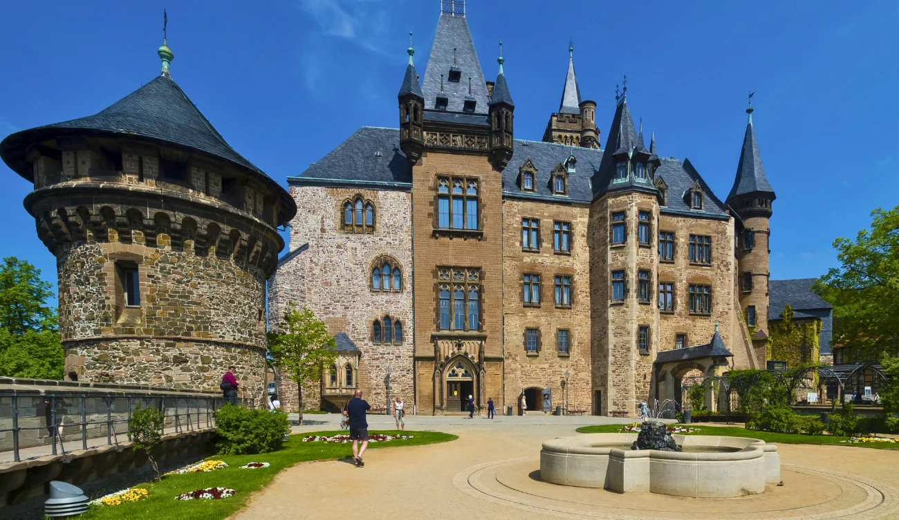 Monumentalny zamek z kamienia, z wysokimi wieżami i ozdobnymi dachami, otoczony starannie zadbanym dziedzińcem z fontanną i kwietnikami. Wokół budynku spacerują turyści, a pod niebieskim niebem panuje pogodna atmosfera.