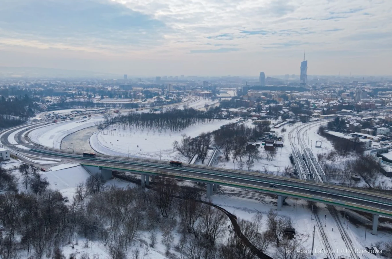 Panorama miasta zimą z widokiem na zaśnieżone ulice, autostradę i tory kolejowe, otoczone drzewami, w tle zabudowania i charakterystyczny wysoki wieżowiec