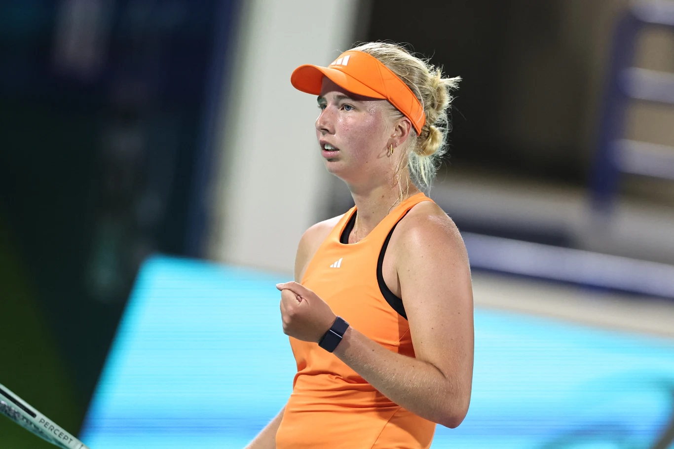 Clara Tauson Kobieta w sportowym stroju w intensywnie pomarańczowym kolorze stoi na korcie tenisowym z rakietą tenisową w dłoni, skupiona podczas meczu, z widoczną opaską na głowie oraz zegarkiem sportowym na nadgarstku.